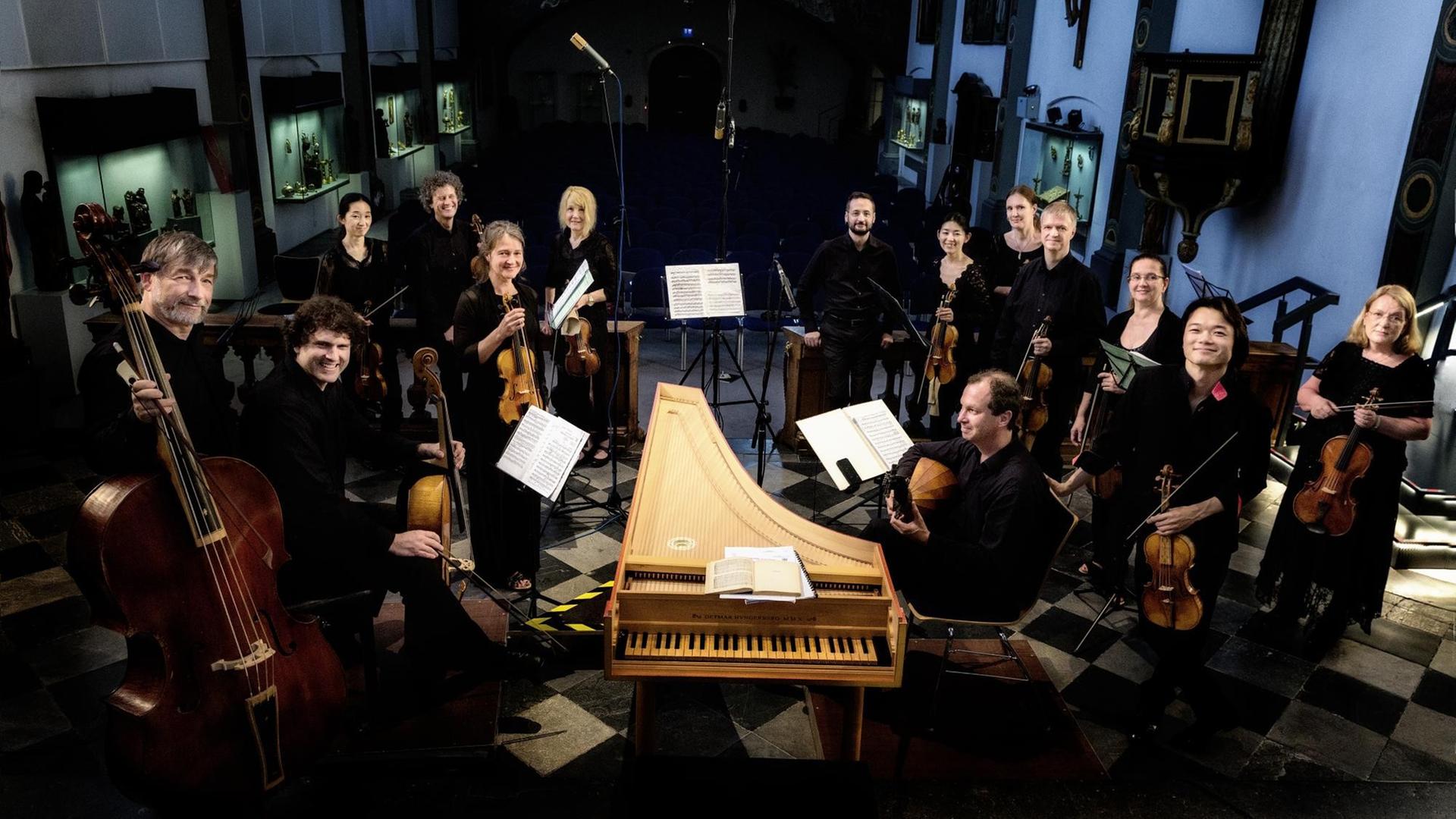 Das Ensemble Concerto Köln steht im Halbkreis um das Cembalo, die Musikerinnen und Musiker halten ihre Instrumente in der Hand, sie tragen schwarze Kleidung und schauen in die Kamera