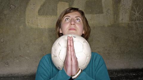 Eine junge Frau mit Fußball und gefalteten Händen hat ihren Blick nach oben gerichtet.