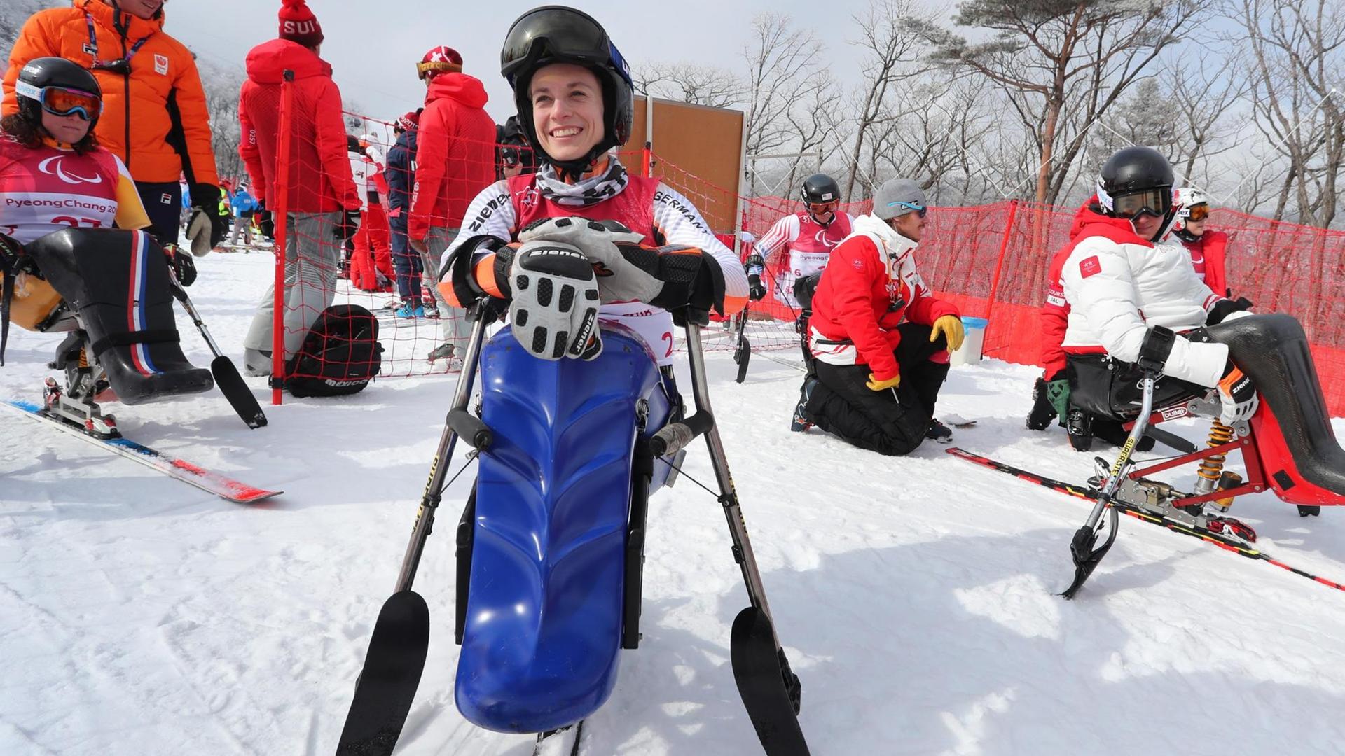Schaffelhuber sitzt in ihrem blauen Monoski und lacht. Dahinter weitere Sportler sowie Betreuer vor einem roten Fangzzaun. Schaffelhuber sitzt in ihrem blauen Monoski und lacht. Dahinter weitere Sportler sowie Betreuer vor einem roten Fangzzaun.
