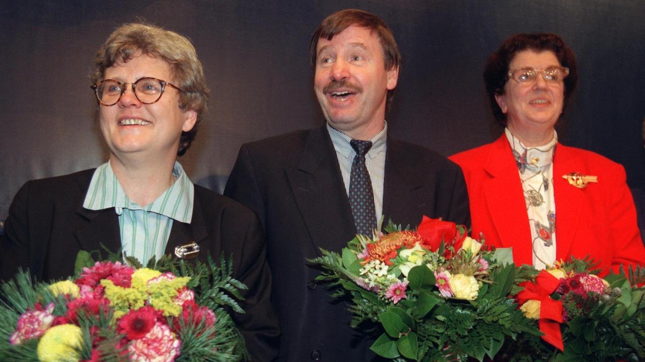 Drei Menschen stehen auf einer Bühne, in den Händen halten sie bunte Blumensträuße. Lore Peschel-Gutzeit steht rechts. 