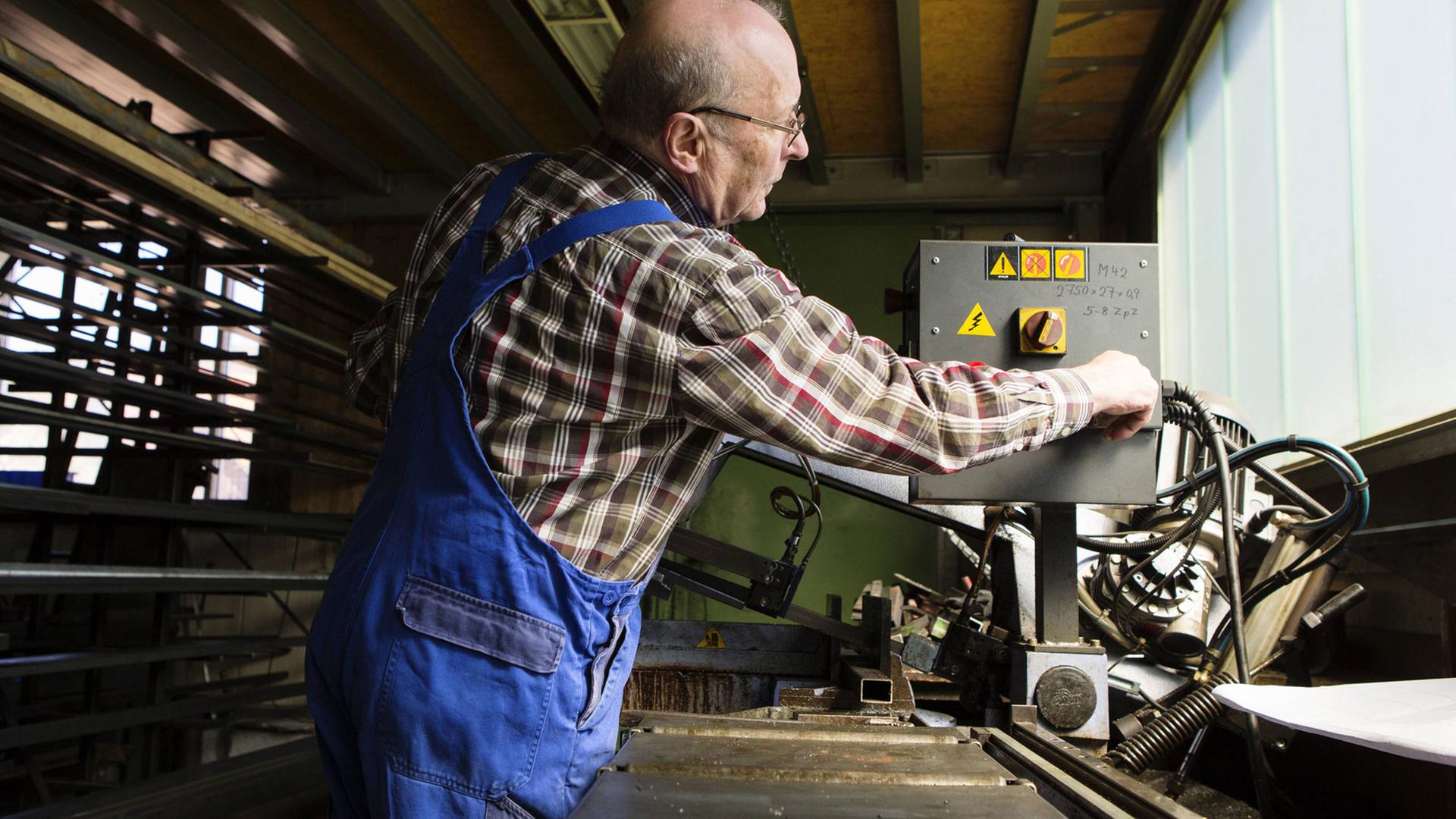 Ein älterer Mann in kariertem Hemd und blauer Arbeitshose bedient eine Maschine in einem mittelständischem Unternehmen Ein älterer Mann in kariertem Hemd und blauer Arbeitshose bedient eine Maschine in einem mittelständischem Unternehmen