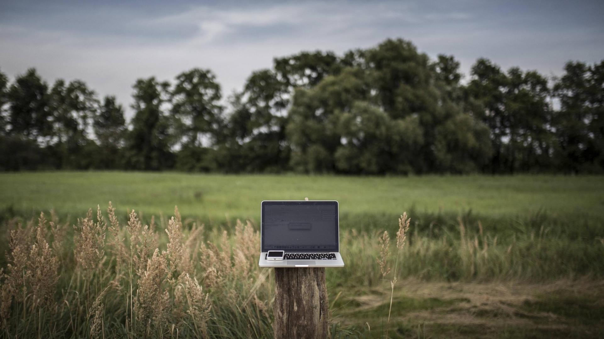 Ein Laptop, mit einem Smartphone darauf, steht auf einem Holzpfeiler in einer grünen Lanschaft. Ein Laptop, mit einem Smartphone darauf, steht auf einem Holzpfeiler in einer grünen Lanschaft.