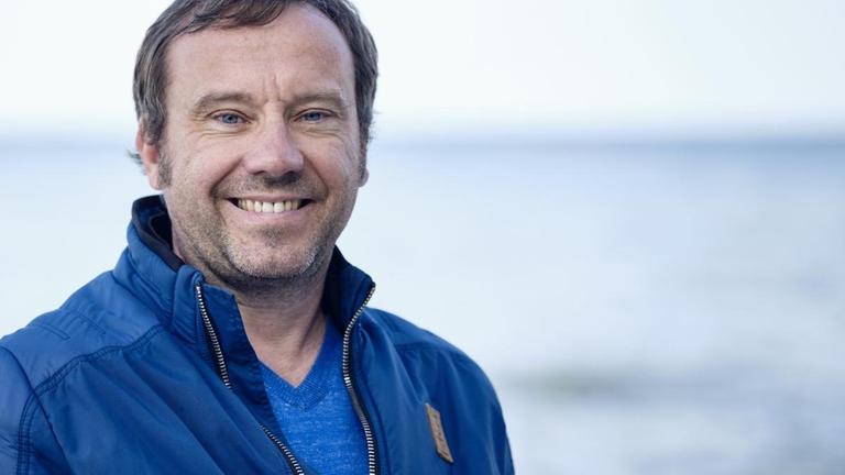 Gregor Sander steht am Meer und lächelt in die Kamera. Gregor Sander steht am Meer und lächelt in die Kamera.