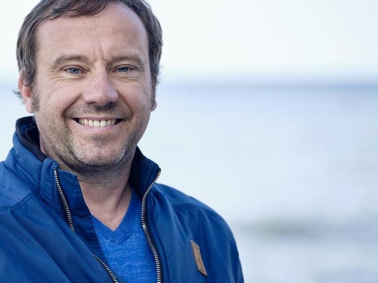 Gregor Sander steht am Meer und lächelt in die Kamera.
