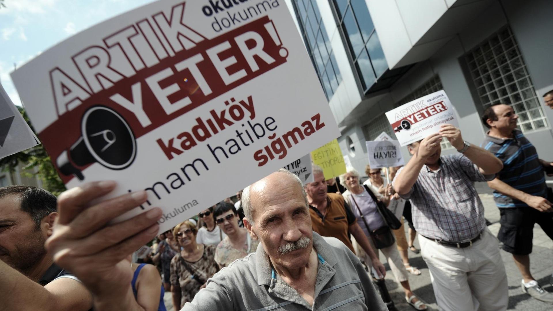 Ein Mann hält ein Protestplakat in die Kamera auf dem auf Türkisch steht: "Genug! Hört auf, unsere Schulen in Imam Hatips zu verwandeln!", im Hintergrund gehen auf einer Straße weitere Demonstranten.