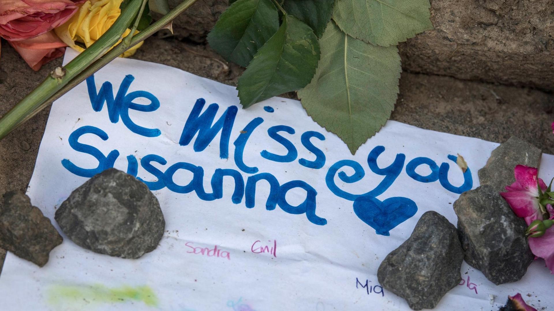 Auf einem weißen Zettel am Boden liegend neben Blumen und Steine steht "We muss you Susanna". Auf einem weißen Zettel am Boden liegend neben Blumen und Steine steht "We muss you Susanna".