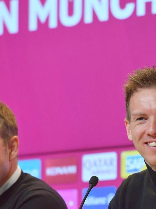 Zwei Männer sitzen in einer Pressekonferenz. Hinter ihnen ist ein magenta farbener Hintergrund auf dem Bayern München steht. Es sind Hansi Flick und sein Nachfolger Julian Nagelsmann.