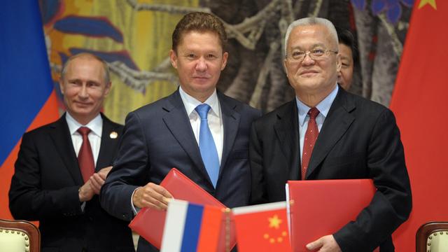 Miller und Zhou posieren mit den Verträgen in roten Mappen in Händen für die Kameras, vor ihnen zwei kleine Flaggen Russlands und Chinas, links hinter ihnen der applaudierende Putin