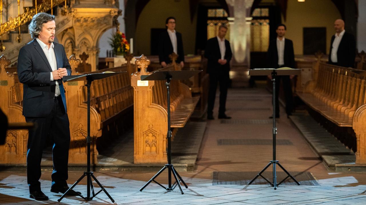 Einer der fünf Sänger des Ensembles stehen im Vordergrund gut beleuchtet vor Kirchenbänken, die im Hintergrund verschwimmen, wo auch die anderen Mitglieder stehen. Einer der fünf Sänger des Ensembles stehen im Vordergrund gut beleuchtet vor Kirchenbänken, die im Hintergrund verschwimmen, wo auch die anderen Mitglieder stehen.
