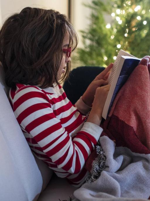Ein Kind sitzt im Schlafanzug auf dem Sofa vor dem Christbaum und liest in einem Buch.