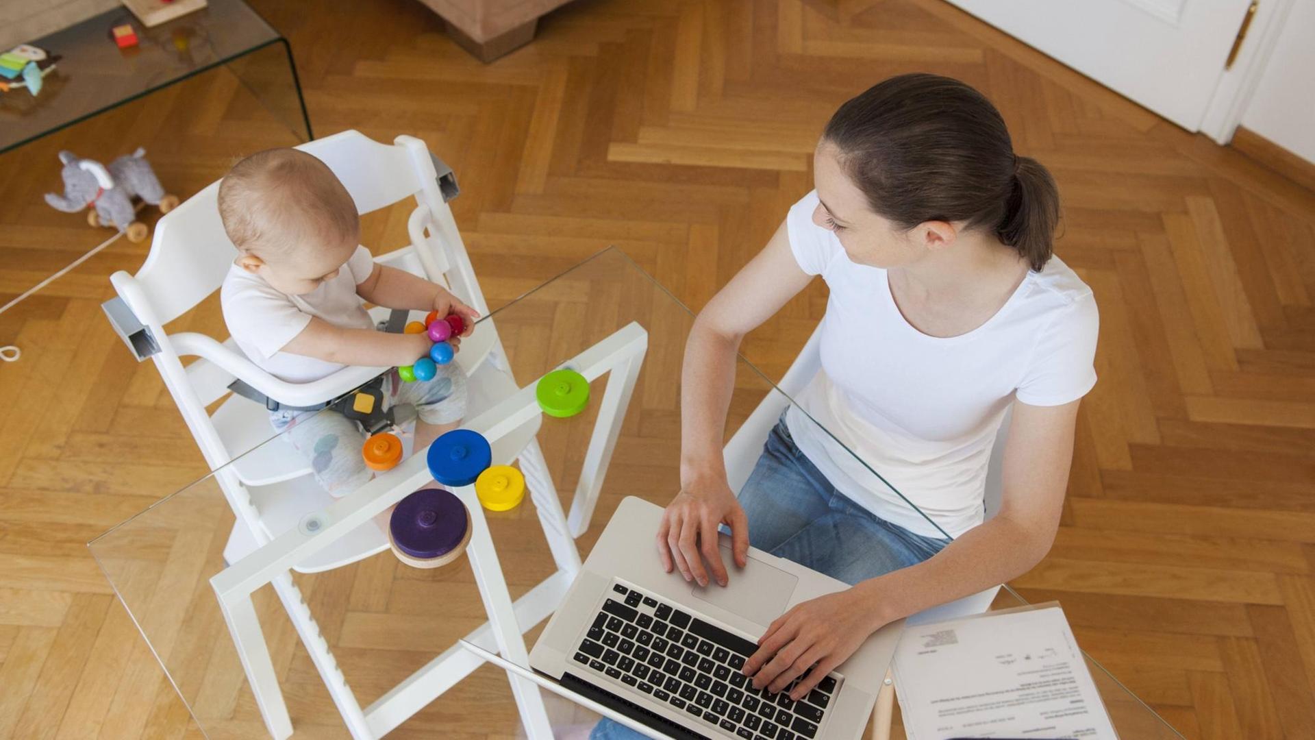 Eine Frau sitzt neben einem ein Baby im Hochstul am Tisch vor einem Laptop