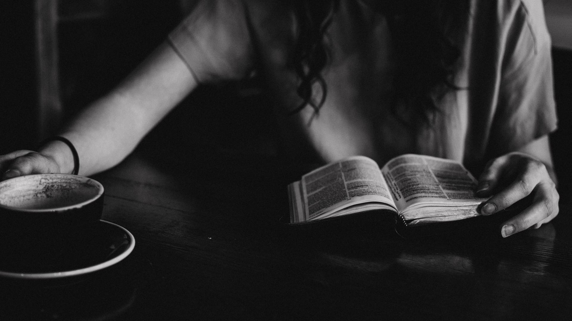 SW-Foto, das die Hände einer Frau zeigt, die in einem Cafe sitzt, und bei einer Tasse Kaffee ein Buch liest.