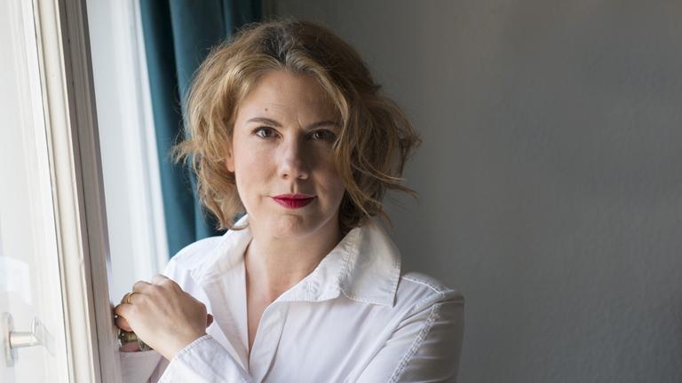 Nina George, im weißen Hemd, mit blondem, schulterlangem Haar und rot geschminkten Lippen, steht an einem Fenster, so dass von links Licht auf ihr Gesicht fällt. Nina George, im weißen Hemd, mit blondem, schulterlangem Haar und rot geschminkten Lippen, steht an einem Fenster, so dass von links Licht auf ihr Gesicht fällt.