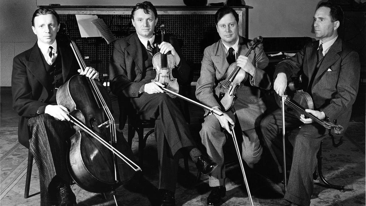 Schwarz-weiß Fotografie. Die vier Mitglieder des Streichquartetts sitzen mit ihren Instrumenten auf Stühlen und schauen ernst in die Kamera. Sie tragen Anzüge in gedeckten Farben, Westen, weiße Hemden und dunkle Schlipse. sie haben kurze dunkle Haare. Schwarz-weiß Fotografie. Die vier Mitglieder des Streichquartetts sitzen mit ihren Instrumenten auf Stühlen und schauen ernst in die Kamera. Sie tragen Anzüge in gedeckten Farben, Westen, weiße Hemden und dunkle Schlipse. sie haben kurze dunkle Haare.