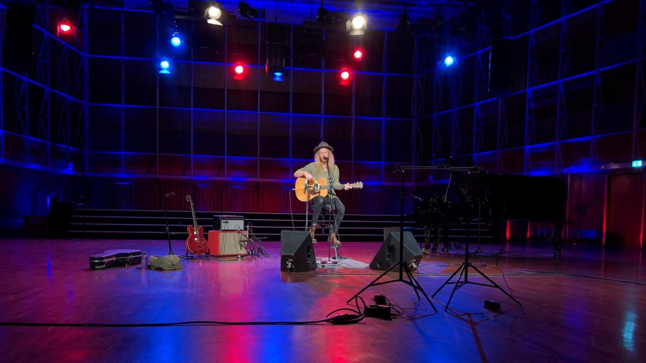 Ein Mann mit langen blonden Haaren und Hut sitzt auf einem Hocker in einem großen Raum. Er hält eine akustische Gitarre in der Hand und ist von mehreren Lautsprechern umgeben. Ein Mann mit langen blonden Haaren und Hut sitzt auf einem Hocker in einem großen Raum. Er hält eine akustische Gitarre in der Hand und ist von mehreren Lautsprechern umgeben.