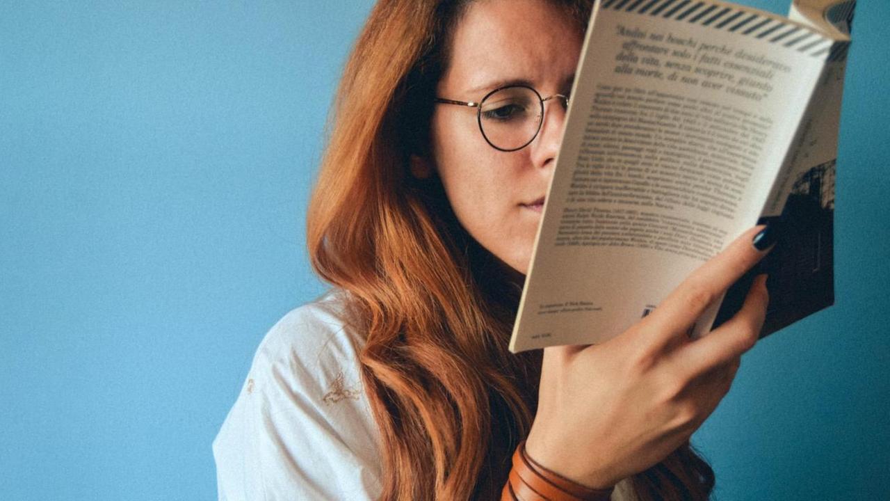 Eine junge Frau mit Brille sitzt vor einer blauen Wand und liest. Eine junge Frau mit Brille sitzt vor einer blauen Wand und liest.