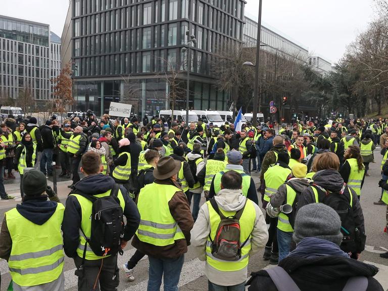 In Paris protestieren Anhänger der sogenannten "Gelbwesten", im Hintergrund ist ein Bürogebäude zu sehen.  