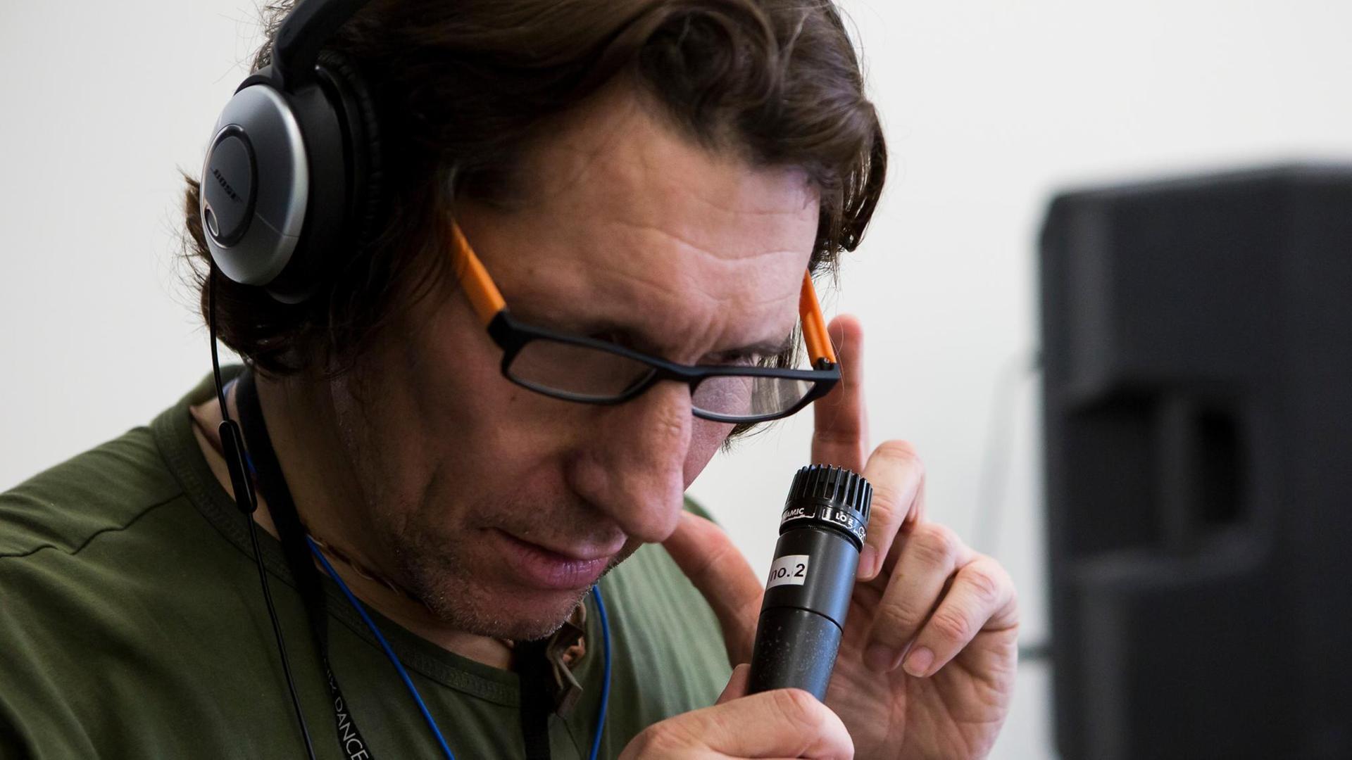 Ein Mann mit Brille trägt Kopfhörer und hält ein Mikrofon in der rechten Hand, sein Blick geht nach unten. Ein Mann mit Brille trägt Kopfhörer und hält ein Mikrofon in der rechten Hand, sein Blick geht nach unten.