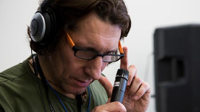 Ein Mann mit Brille trägt Kopfhörer und hält ein Mikrofon in der rechten Hand, sein Blick geht nach unten.