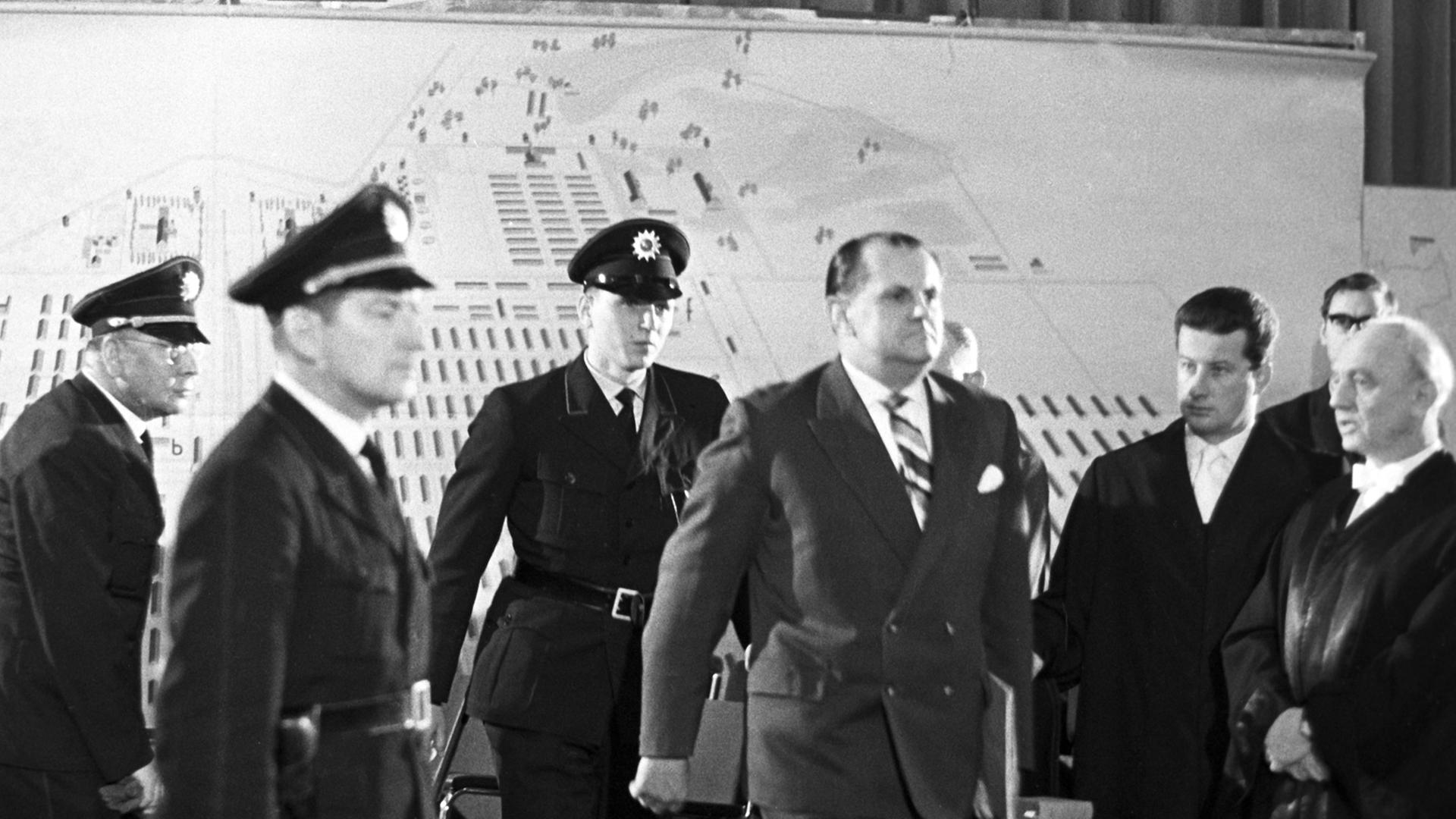 Der Angeklagte Oswald Kaduk wird von drei Polizisten in einen Gerichtssaal geführt. Der Angeklagte Oswald Kaduk wird von drei Polizisten in einen Gerichtssaal geführt.