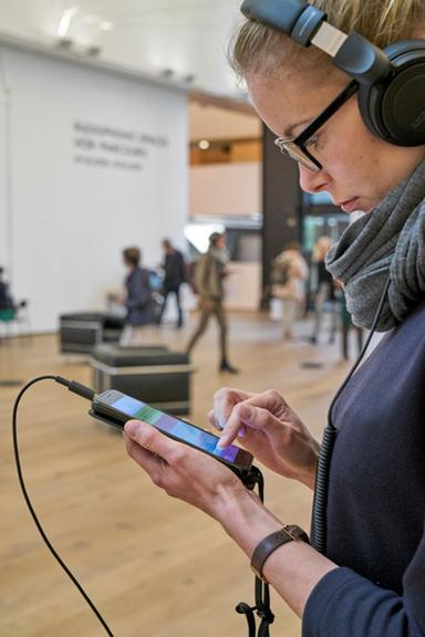 Installationsansicht "Radiophonic Spaces" im Museum Tinguely, Basel. Eine Frau steht im Profil in einem Ausstellungsraum, trägt eine Brille, hat einen Kopfhörer auf und schaut auf ihr Smartphone
