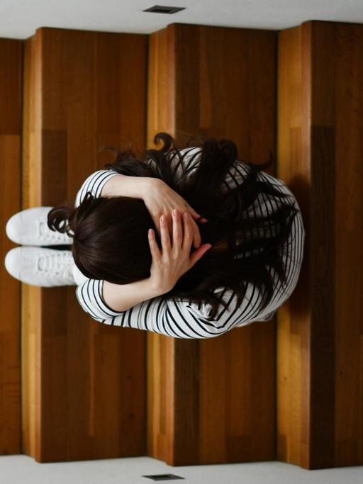 Illustrationsfoto zum Thema Depressionen: Eine Frau kauert auf einer Treppe. Das Foto zeigt sie von oben. Sie hat die Arme um ihren Körper geschlungen. Foto: Frank May/picture alliance (model released) | Verwendung weltweit