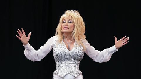 Dolly Parton auf einem Foto vom 29. Juni 2014. Sie träge weiß, eng anliegende Kleidung und breitet die Arme aus. Ihre Haare sind platinblond.