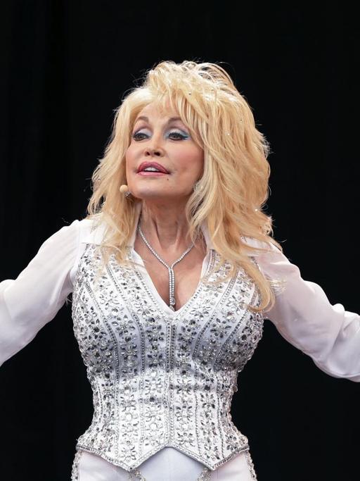 Dolly Parton auf einem Foto vom 29. Juni 2014. Sie träge weiß, eng anliegende Kleidung und breitet die Arme aus. Ihre Haare sind platinblond.