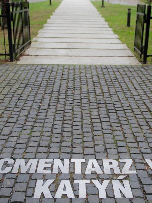 Blick auf einen Friedhof durch das Eingangstor, auf dem Boden stehen die Worte: Polski Cmentarz Wojenny Katyn