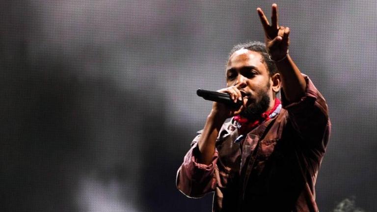 Kendrick Lamar beim Panorama Music Festival 2016 in New York. Der Musiker steht vor dunklem Hintergrund und macht mit einer Hand das Peace-Zeichen.