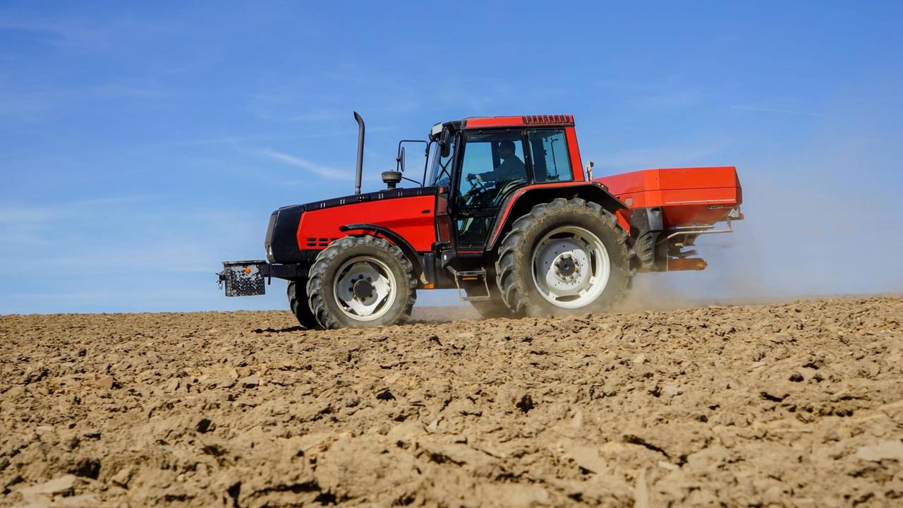 Ein Landwirt düngt ein unbestelltes Feld im Frühjahr mit Mineraldünger, der rote Trecker fährt über den Boden und wirbelt Staub auf. Ein Landwirt düngt ein unbestelltes Feld im Frühjahr mit Mineraldünger, der rote Trecker fährt über den Boden und wirbelt Staub auf.