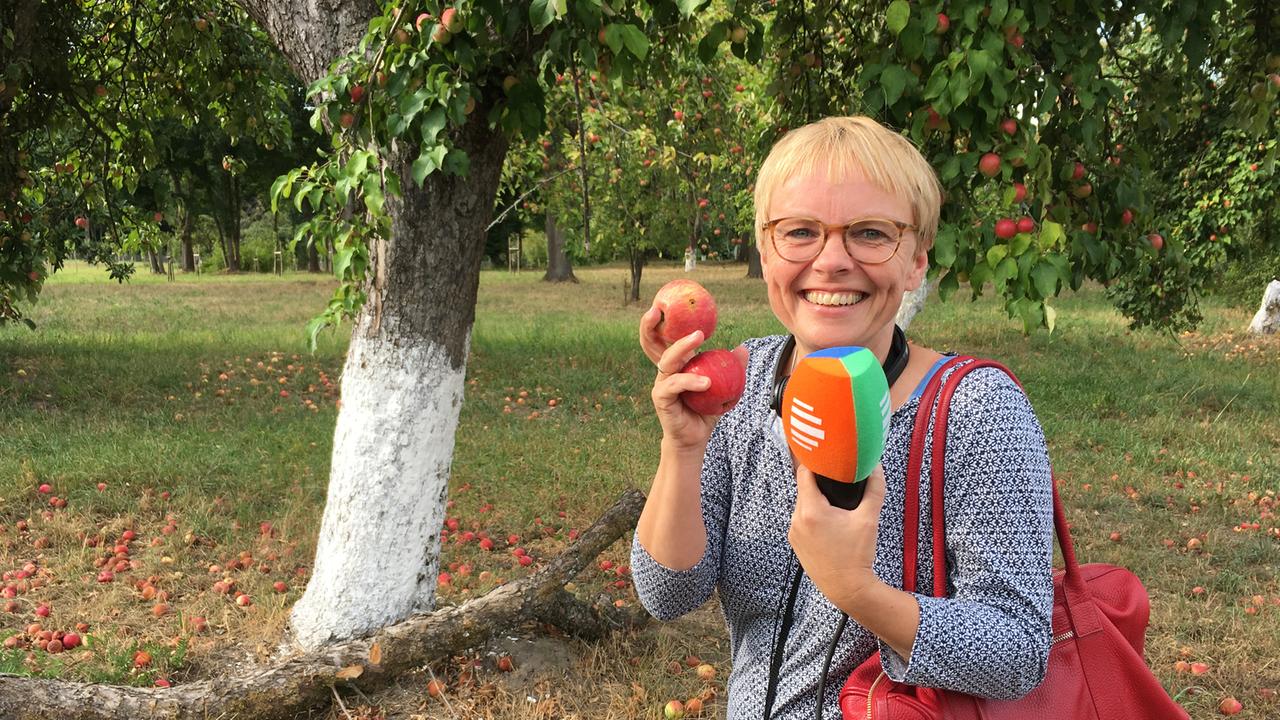 Margarete Wohlan steht im Park, hält Äpfel in der Hand und lächelt