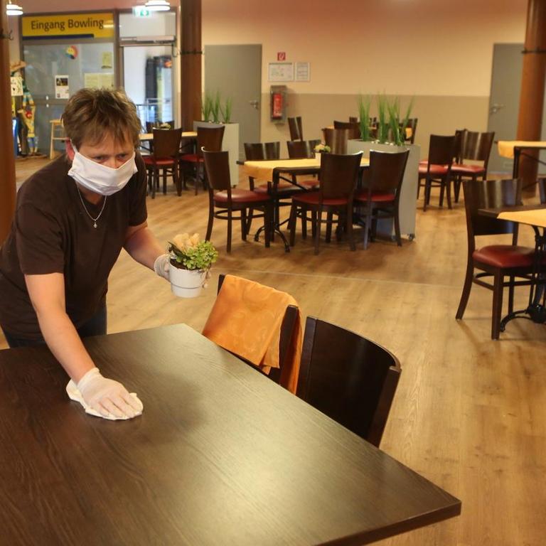 13.05.2020, Sachsen-Anhalt, Wernigerode: Eine Mitarbeiterin mit Mund-Nase-Schutz und einem Reinigungstuch in der Hand beugt sich über einen Restauranttisch.