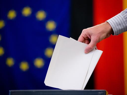 Ein Wähler gibt am 25.05.2014 in Berlin-Mitte seinen Stimmzettel für die Europawahl und den Volksentscheid für das Tempelhofer Feld ab. Im Hintergrund sind eine Europa- und eine Deutschlandfahne zu sehen.