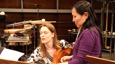 Zwei Frau schauen auf einen Notenständer. Die rechte Frau steht, die linke sitzt und hat ein Cello in der linken Hand. Im Hintergrund stehen weitere Instrumente