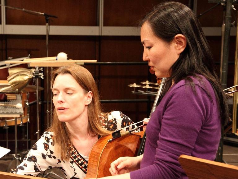 Zwei Frau schauen auf einen Notenständer. Die rechte Frau steht, die linke sitzt und hat ein Cello in der linken Hand. Im Hintergrund stehen weitere Instrumente