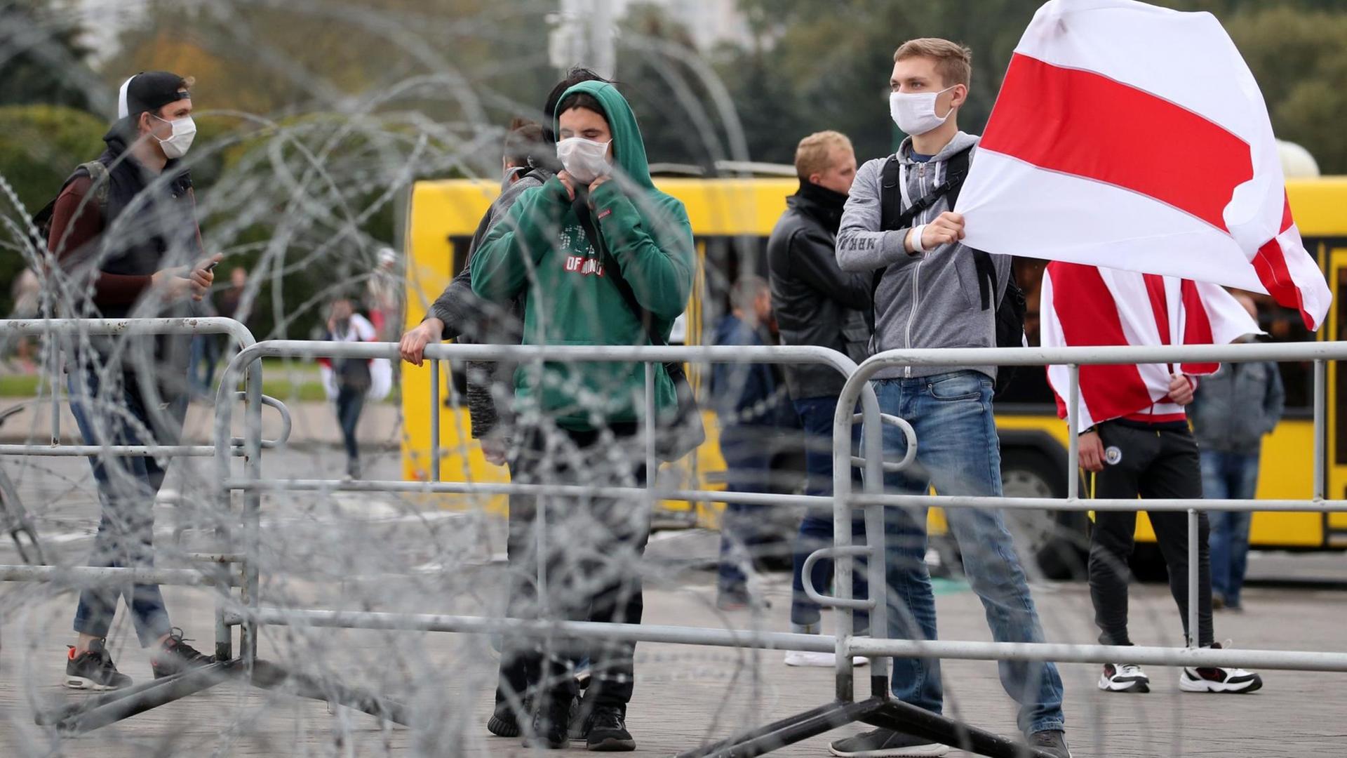 Ein junger Mann hält eine weiß-rote Flagge von Belarus hoch. Er steht hinter einer Absperrung und Stachel-Draht. Ein junger Mann hält eine weiß-rote Flagge von Belarus hoch. Er steht hinter einer Absperrung und Stachel-Draht.
