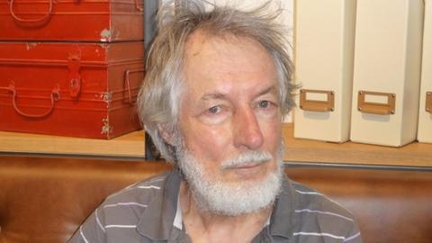 Ein Mann mit Bart und grauen Haaren sitzt von einem Bord mit Ordnern