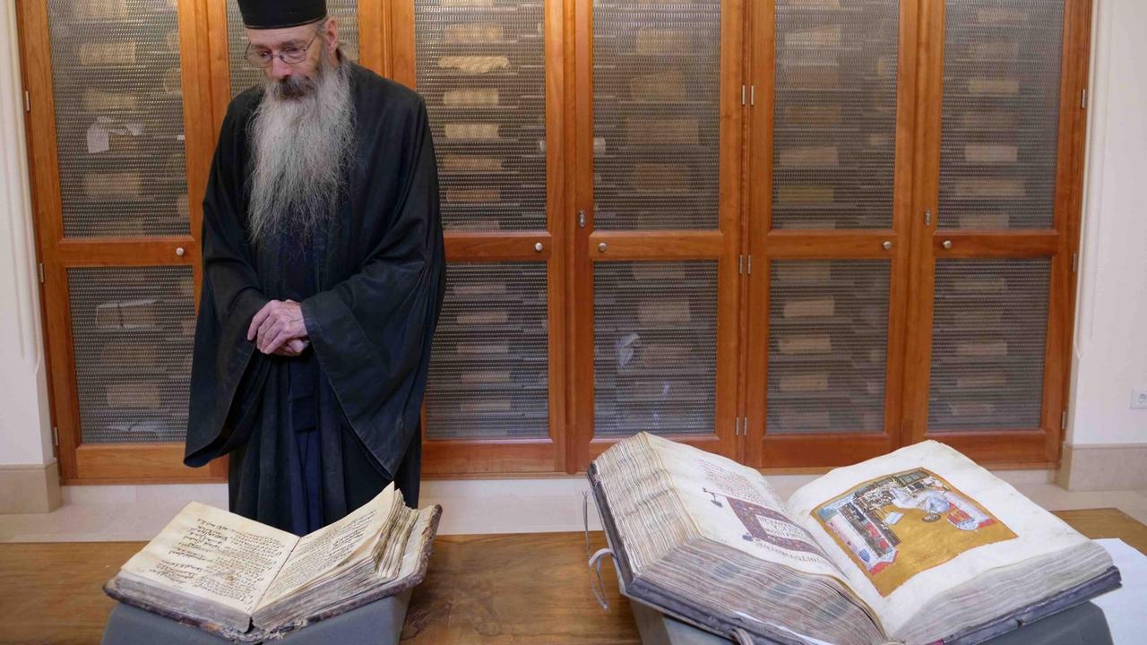 Ein Mann mit einem langen weißen Bart in schwarzer Kutte und mit einer schwarzen Kappe steht mit andächtig übereinander gelegten Händen vor einem Tisch, auf dem zwei große aufgeschlagene Bücher liegen, eines davon mit religiösen Illustrationen. Hinter ihm: die Türen eines Aktenschranks mit hellen Holzrahmen. Ein Mann mit einem langen weißen Bart in schwarzer Kutte und mit einer schwarzen Kappe steht mit andächtig übereinander gelegten Händen vor einem Tisch, auf dem zwei große aufgeschlagene Bücher liegen, eines davon mit religiösen Illustrationen. Hinter ihm: die Türen eines Aktenschranks mit hellen Holzrahmen.