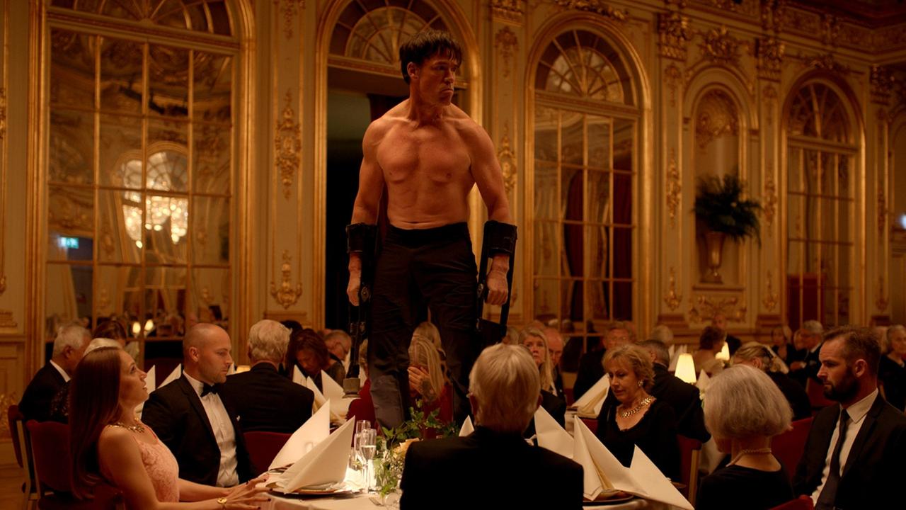 Ein einen Affen mimender Künstler (Terry Notary) steht auf einem Tisch, die chic gekleideten Gäste blicken erschrocken zu ihm auf Ein einen Affen mimender Künstler (Terry Notary) steht auf einem Tisch, die chic gekleideten Gäste blicken erschrocken zu ihm auf