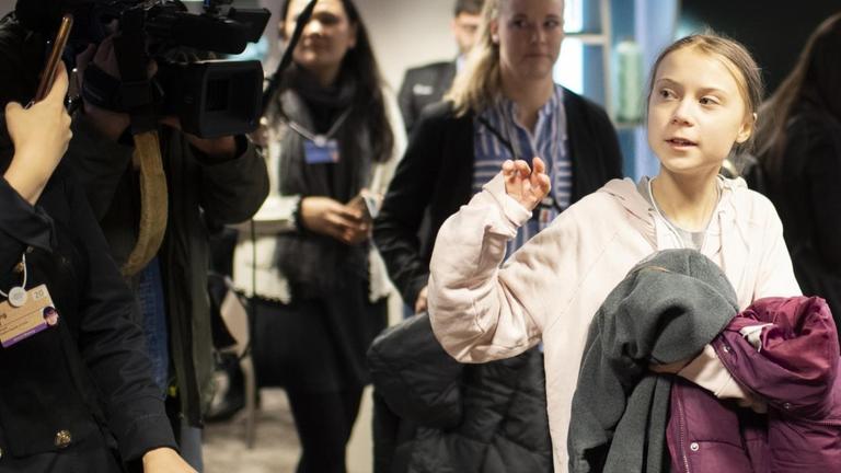 Greta Thunberg läuft umringt von Journalisten und Medientechnikern durch einen Flur und trägt ihre Jacke über den Arm geschlagen. Greta Thunberg läuft umringt von Journalisten und Medientechnikern durch einen Flur und trägt ihre Jacke über den Arm geschlagen.