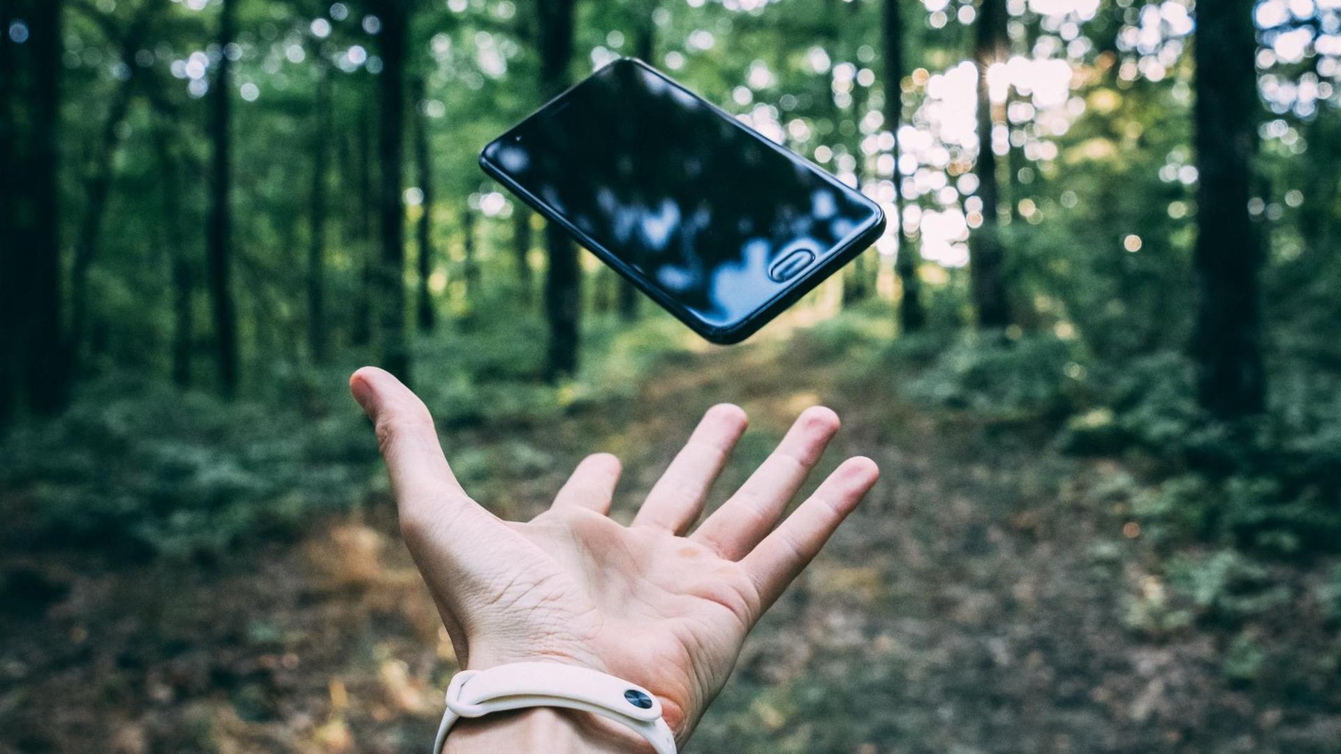 Ein Handy fliegt durch die Luft, eine Hand versucht es aufzufangen. Im Hintergrund Wald. Ein Handy fliegt durch die Luft, eine Hand versucht es aufzufangen. Im Hintergrund Wald.