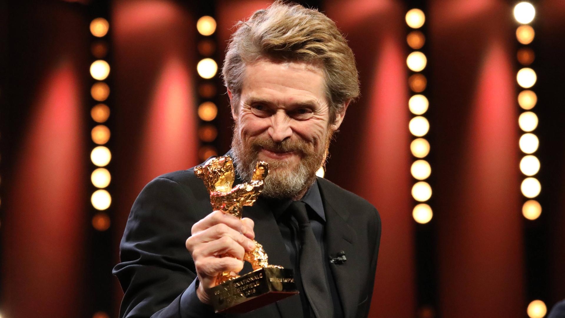 Der Schau-Spieler Willem Dafoe hält einen kleinen goldenen Bären in der Hand. Der Schau-Spieler Willem Dafoe hält einen kleinen goldenen Bären in der Hand.