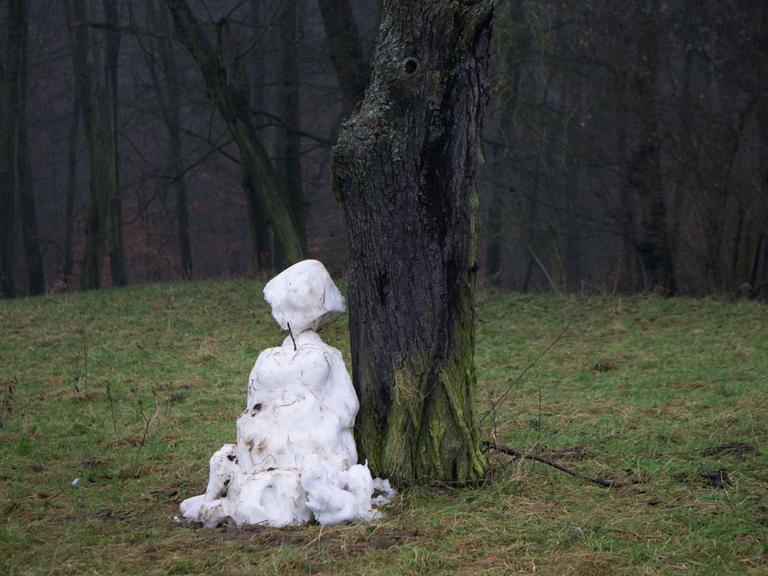 Ein dahinschmelzender Schneemann steht an einem Waldrand auf einer grünen Wiese.