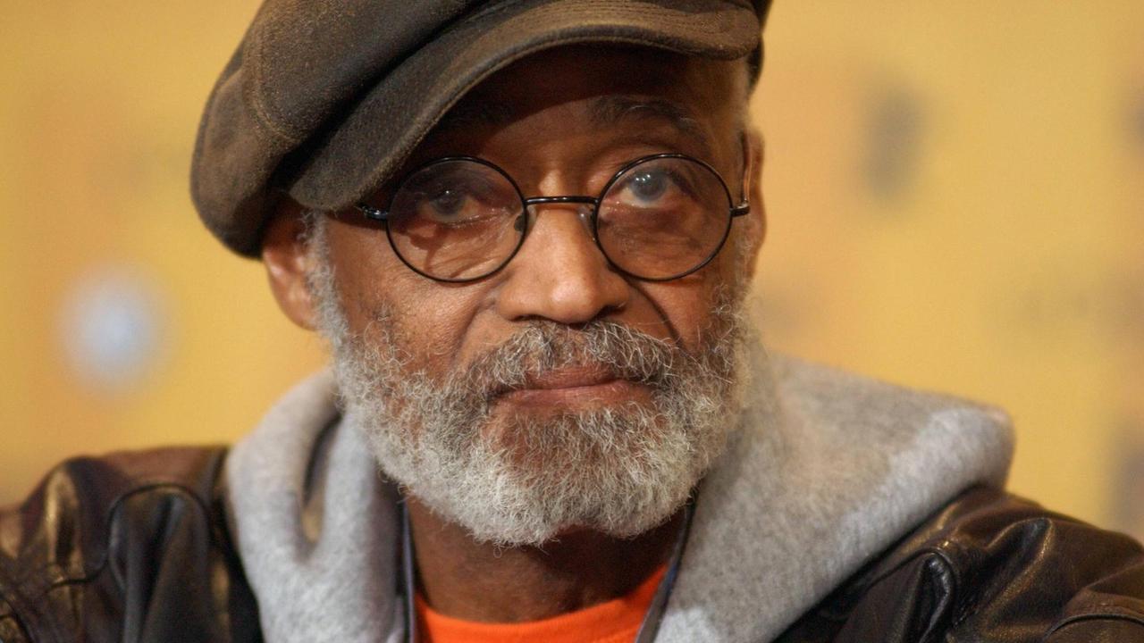 Porträt von Melvin van Peebles. Er trägt seine charakteristische Brille mit runden Gläsern, eine Schiebermütze, Lederjacke und einen grauen Kapuzenpullover. Porträt von Melvin van Peebles. Er trägt seine charakteristische Brille mit runden Gläsern, eine Schiebermütze, Lederjacke und einen grauen Kapuzenpullover.