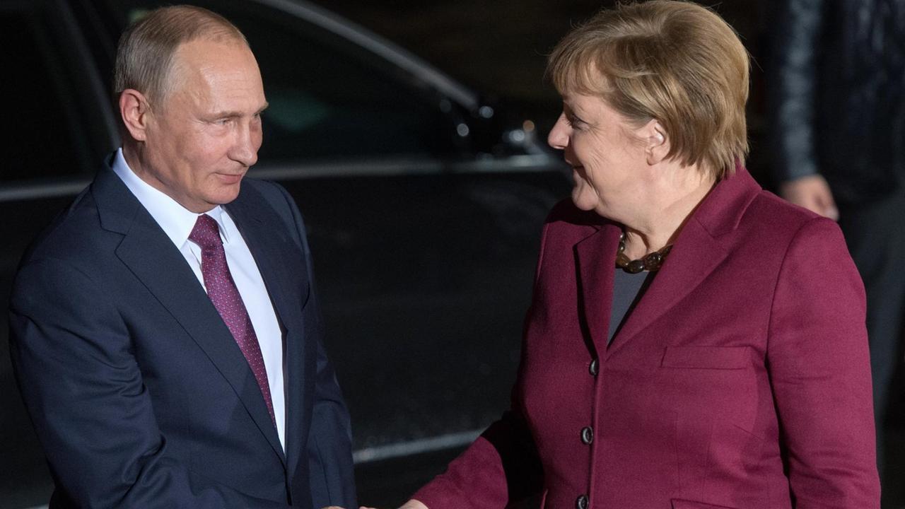Merkel und Putin geben sich vor einem schwarzen Auto die Hand. Beide lächeln. Merkel und Putin geben sich vor einem schwarzen Auto die Hand. Beide lächeln.