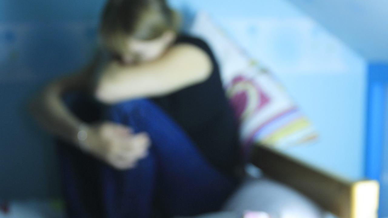 Eine verzweifelte junge Frau hockt auf einem Bett. Im Vordergrund: Tabletten. 
