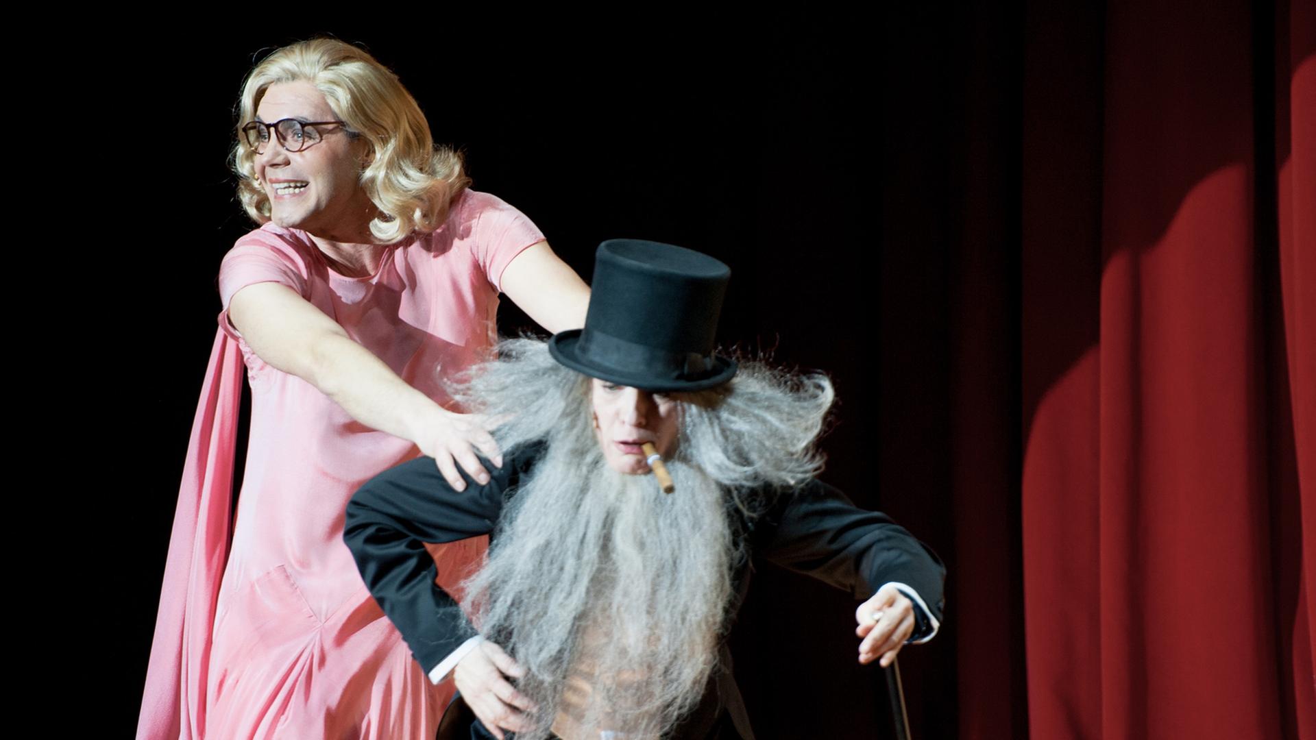 Max Hopp in rosa Kleid mit blonder Perücke und Brille, stützt sich auf Dagmar Manzel in schwarzem Anzug mit grauem langem Bart. Max Hopp in rosa Kleid mit blonder Perücke und Brille, stützt sich auf Dagmar Manzel in schwarzem Anzug mit grauem langem Bart.