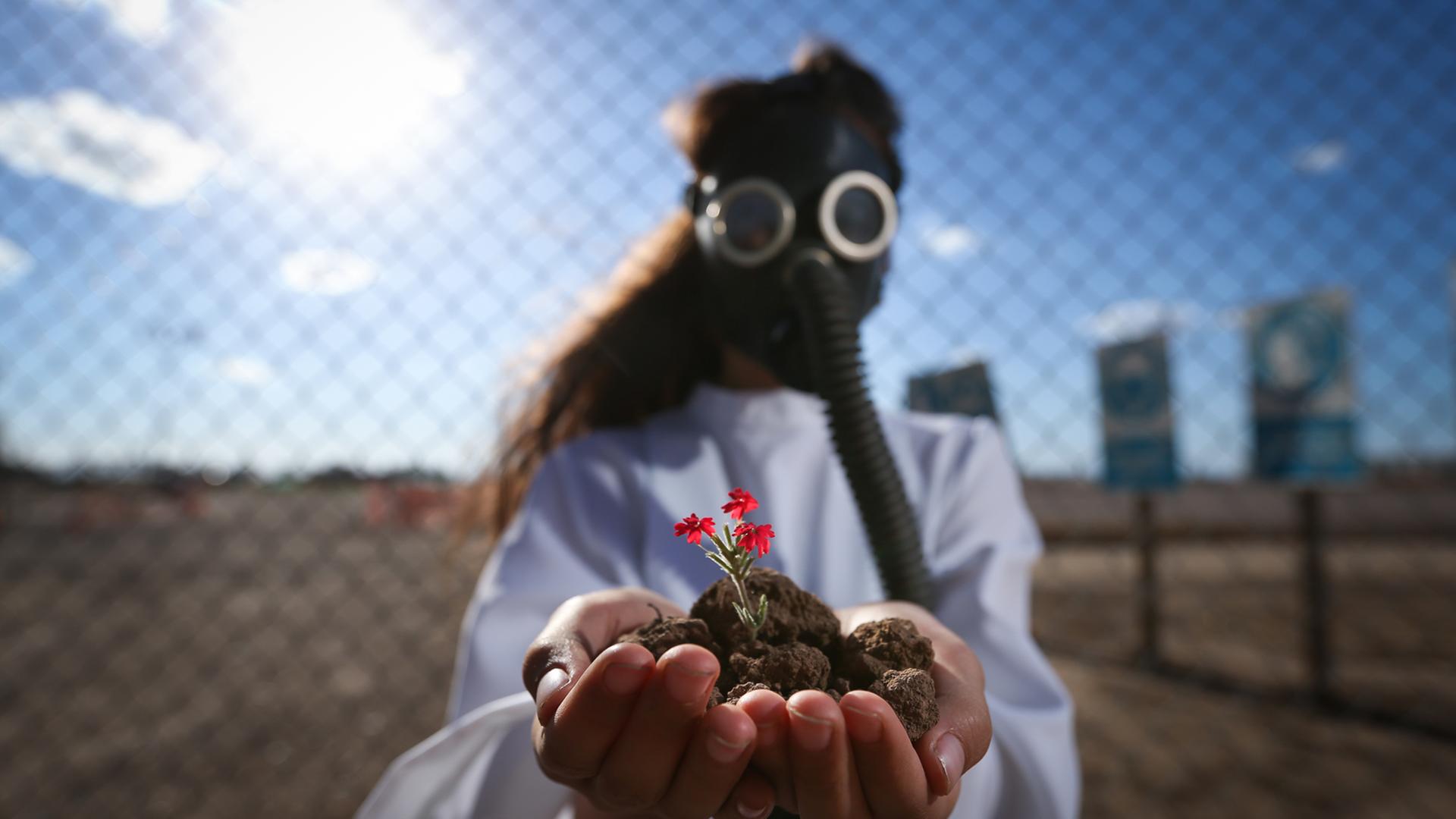 Ein Mädchen vor einem Maschendrahtzaun in einer kargen Landschaft, mit einer Gasmaske und einem weißen Hemd, hält in den göffneten Händen Erde mit einer kleinen rot blühenden Pflanze in die Kamera. Ein Mädchen vor einem Maschendrahtzaun in einer kargen Landschaft, mit einer Gasmaske und einem weißen Hemd, hält in den göffneten Händen Erde mit einer kleinen rot blühenden Pflanze in die Kamera.