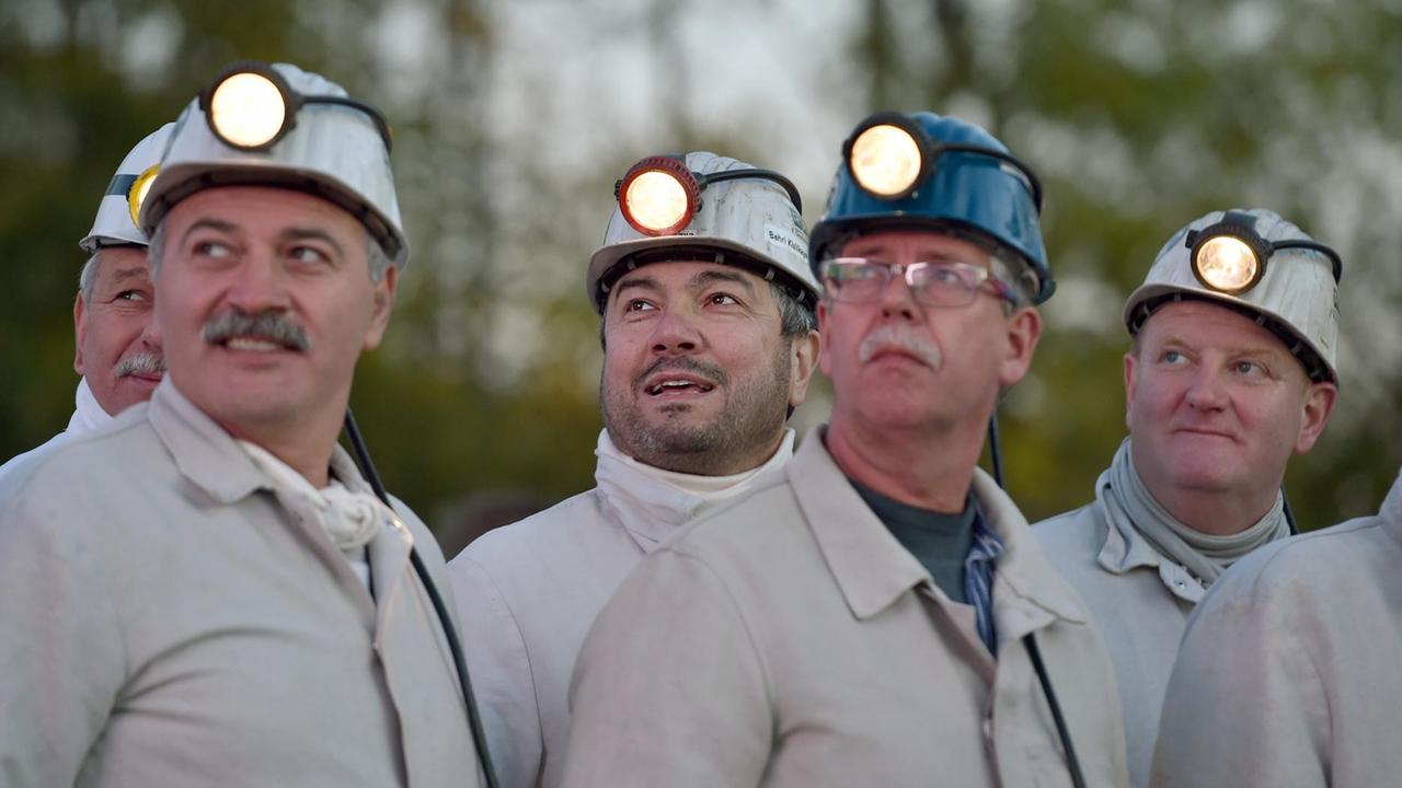 Sechs ältere Bergleute in grauen Arbeitsanzügen und mit Helmen mit Lampen blicken - teils lächelnd - nach rechts oben.